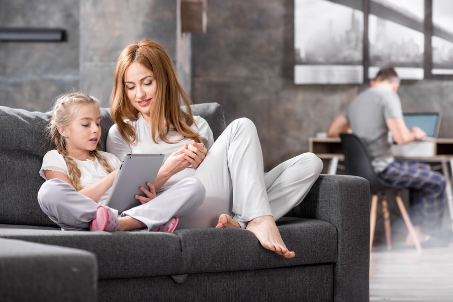 Woman and child sit on a sofa looking at a tablet while man works at a laptop in the background.